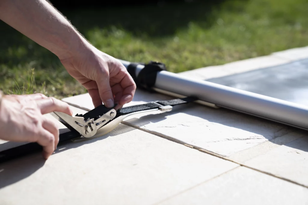 Instalation d'un cliquet en inox robuste pour fixer une sangle de tension sur une couverture à barres de piscine, assurant une fixation stable et sécurisée.